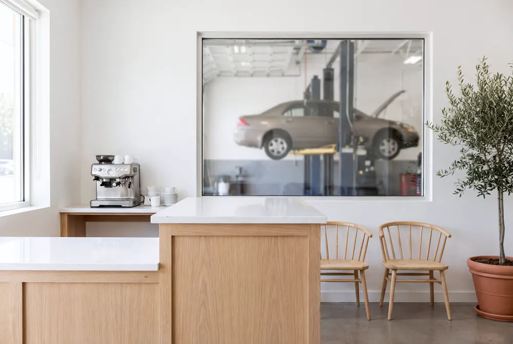 The front counter in pale oak and white quartz, an espresso machine and cups, two wooden chairs and an olive tree, with a window through to a car on a lift.