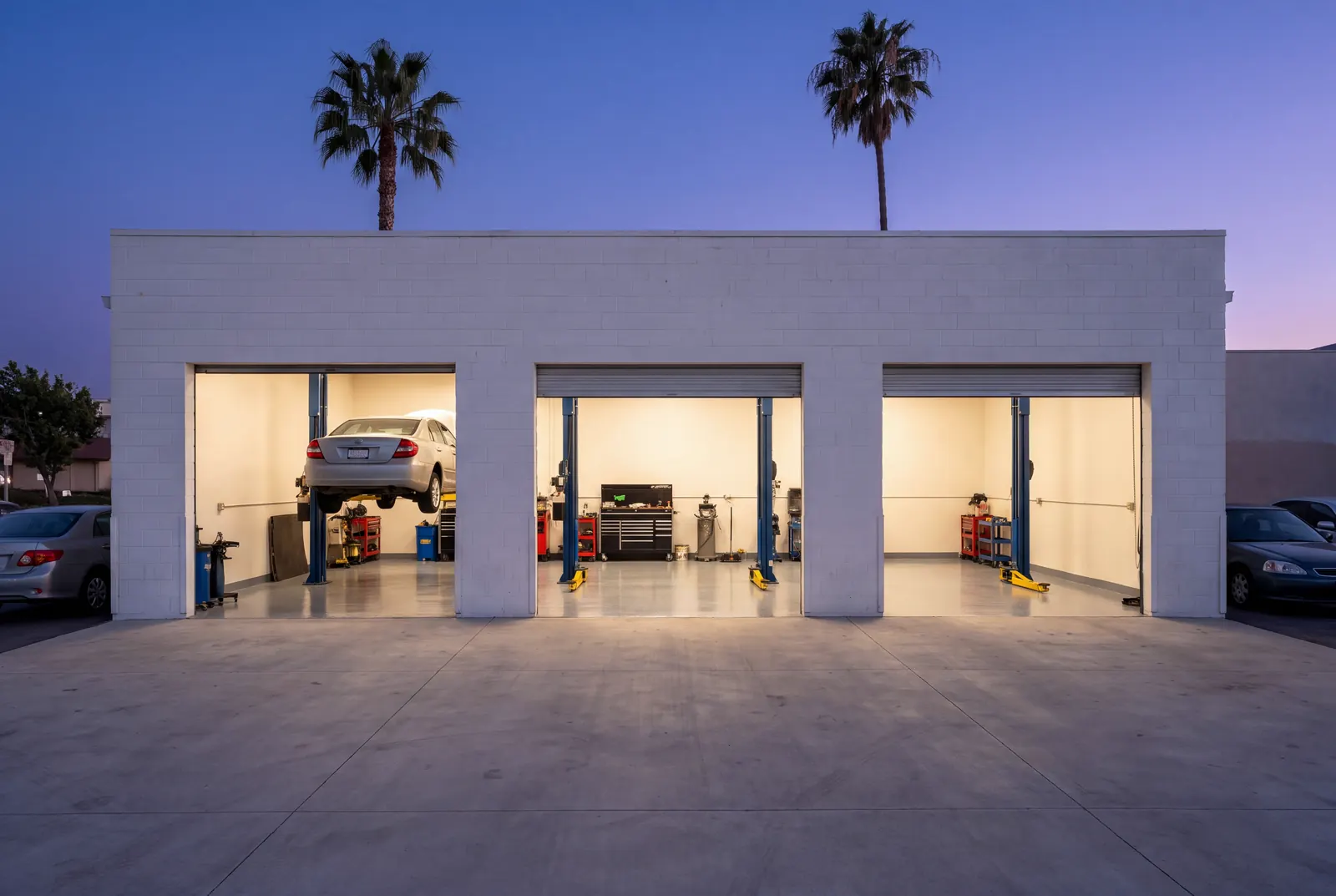The shop from across the street at blue hour: a white block building with three roll-up doors open, the bays lit warm inside, a car on a lift, two palm trees against a deep blue sky.