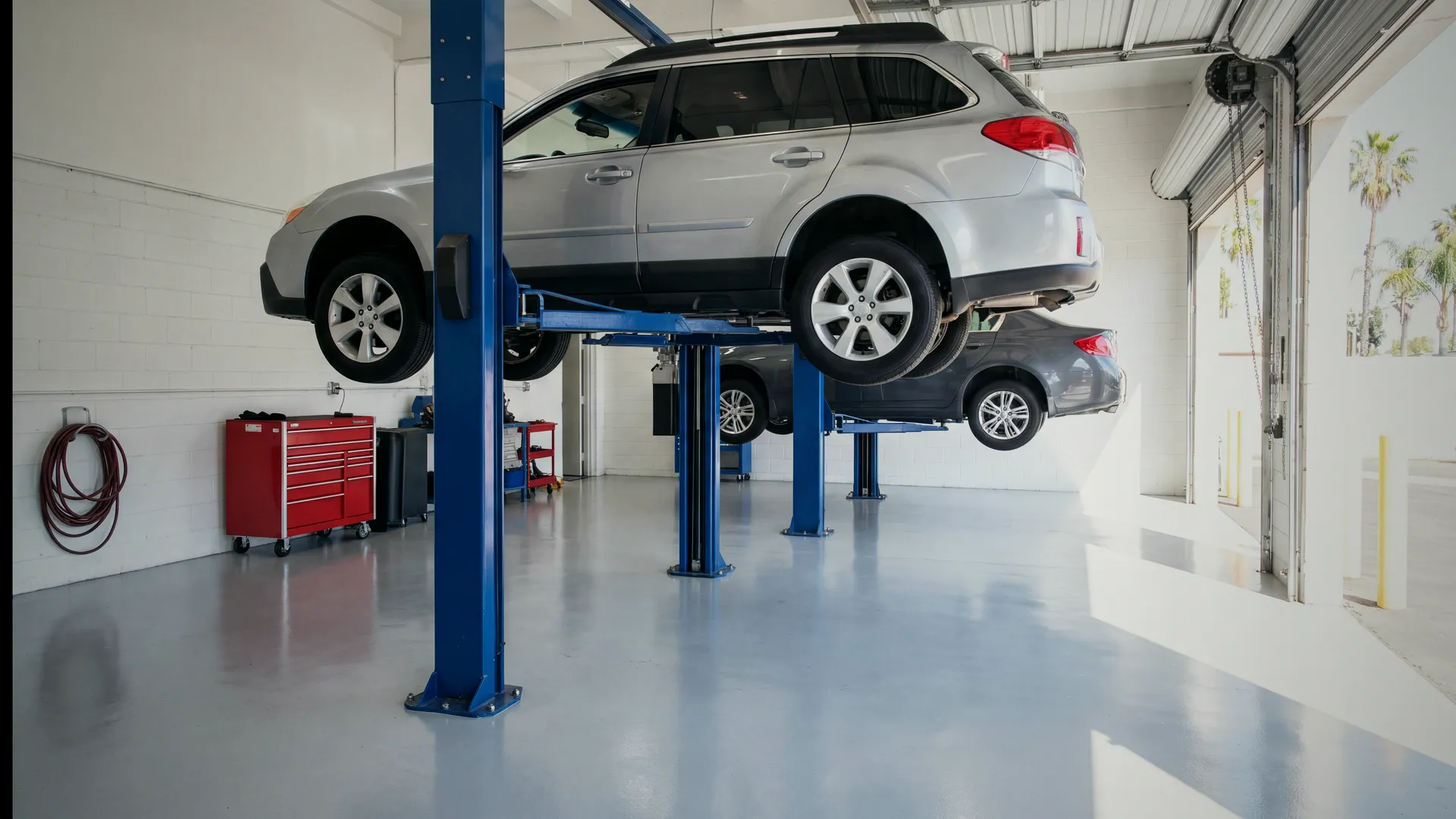 Inside the shop on a weekday morning: a silver Subaru Outback raised on a two-post lift, a grey sedan on the lift behind it, and the roll-up door open to palm trees and daylight.
