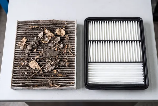 The old cabin air filter, clogged grey-brown with dust and dry leaves, next to a clean new white filter.