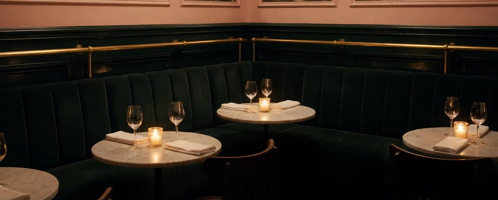The dining room's corner banquette in bottle-green velvet under a brass rail, small marble tables set with candles, wine glasses and white napkins, blush wall above