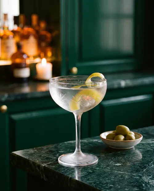 A frosted, cut-glass coupe of gin martini with a long lemon twist on green marble, a small dish of olives beside it