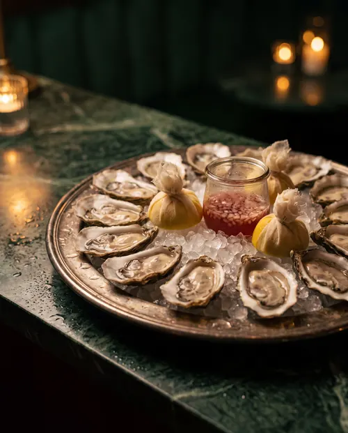 A dozen freshly shucked oysters on crushed ice on a silver platter, with lemon halves tied in muslin and a jar of shallot mignonette