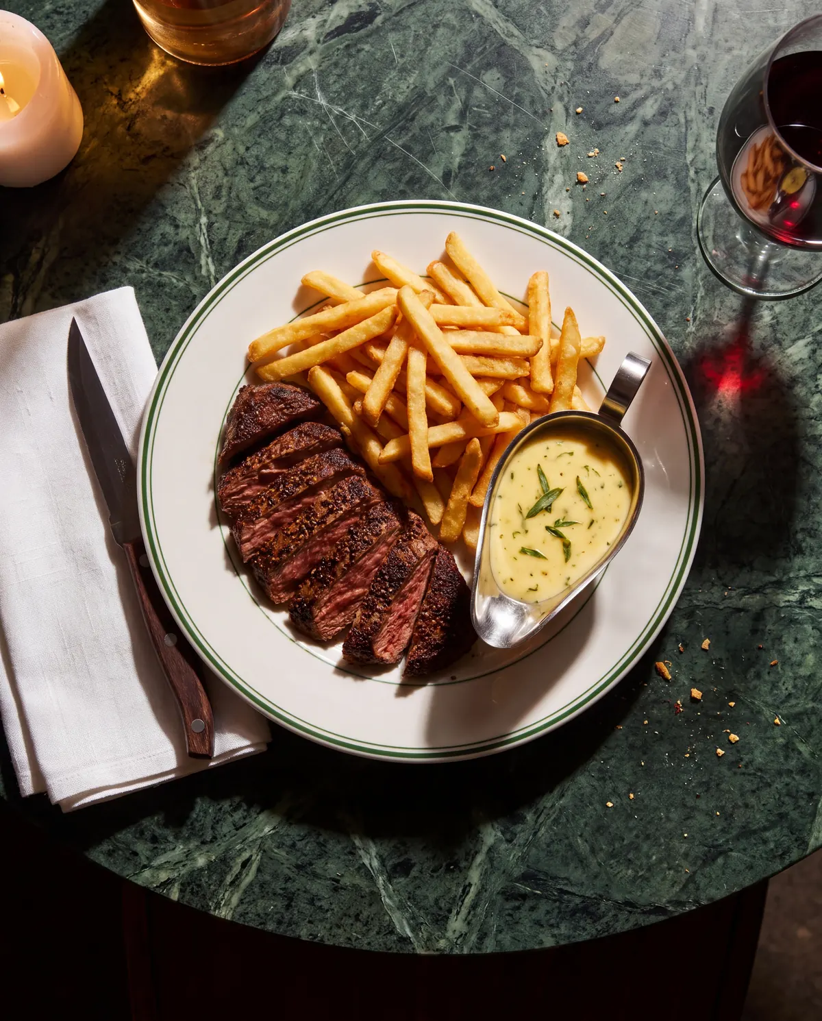 Overhead view of sliced medium-rare bavette with a peppered crust, a heap of frites and a silver boat of béarnaise on a green-rimmed plate, on green marble beside a candle and a glass of red