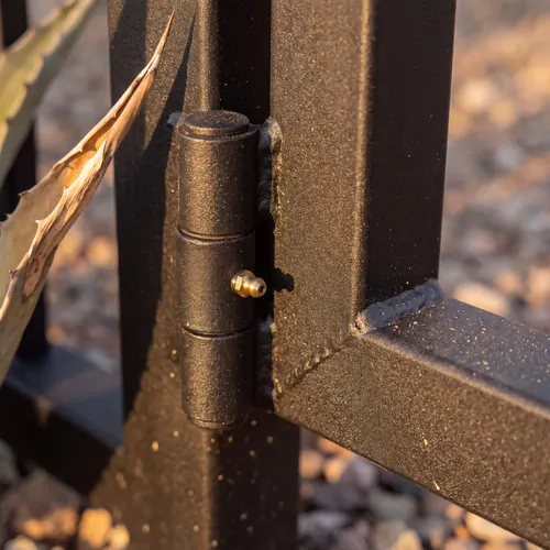 Close-up of a greaseable barrel hinge welded between a black powder-coated gate frame and its square post, lit by low sun.