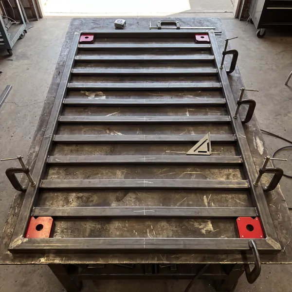 Looking straight down at a gate frame tacked together on the welding table, with magnetic squares in the corners, C-clamps on the sides and chalk marks on each slat.