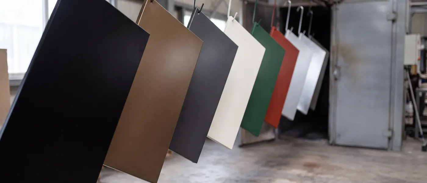 Powder-coated steel sample panels in black, bronze, grey, off-white, green, red and silver hanging from hooks in front of the curing oven.