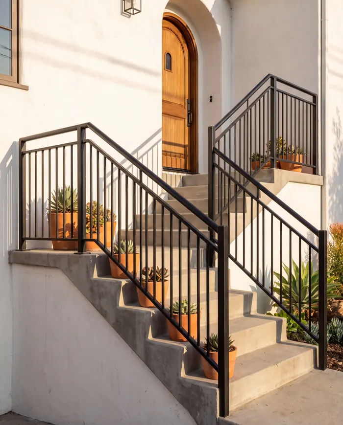 Black steel handrails and pickets running up both sides of a concrete entry stair to an arched wooden front door, terracotta pots on the steps.